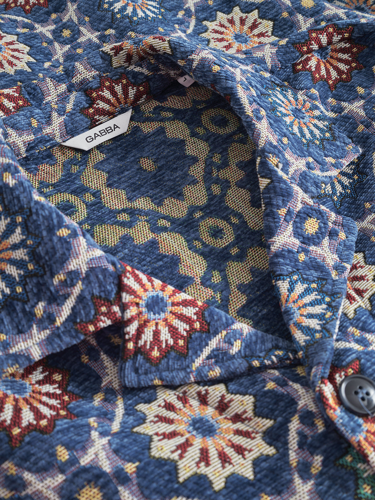 Close-up of the GABsimone Nava LS - Multi Pattern shirt by Gabba, featuring textured jacquard fabric in blue with intricate, colorful floral and geometric designs. A visible white tag inside the collar displays the Gabba brand name.