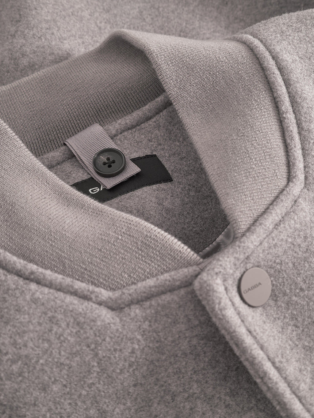 Close-up of the Orta Wox - Sand bomber jacket by Gabba, highlighting the collar, a buttoned fabric tag, and a metal snap fastener engraved with “GABBA.” The light grey wool blend is soft, textured, breathable, and stylish.