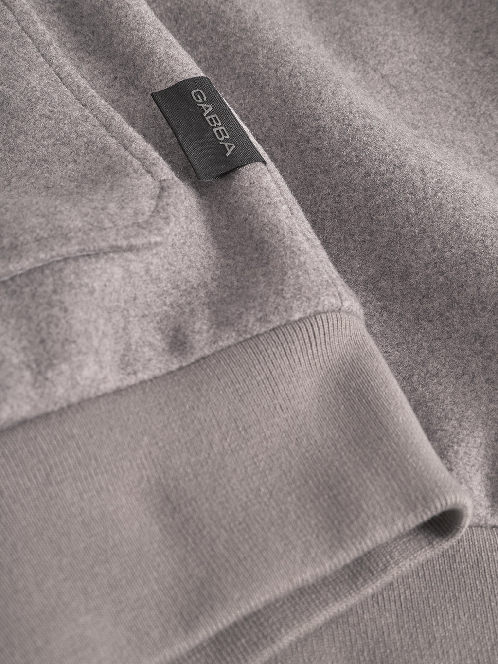 Close-up of the Orta Wox - Sand sweatshirt by Gabba, showing light grey breathable fabric, soft texture, ribbed cuff, and a black label with the Gabba brand name in white.