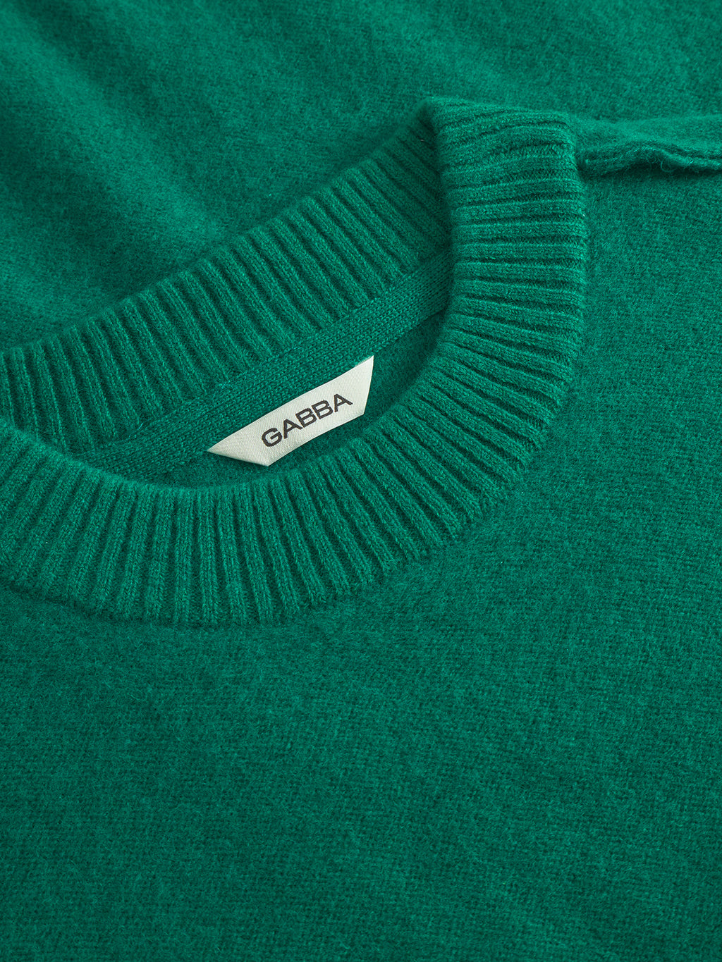 Close-up of the Tokota Aico LS sweater in green by Gabba, featuring a ribbed crew neckline and a white collar tag with 'Gabba' in black, showcasing its fine merino wool quality.
