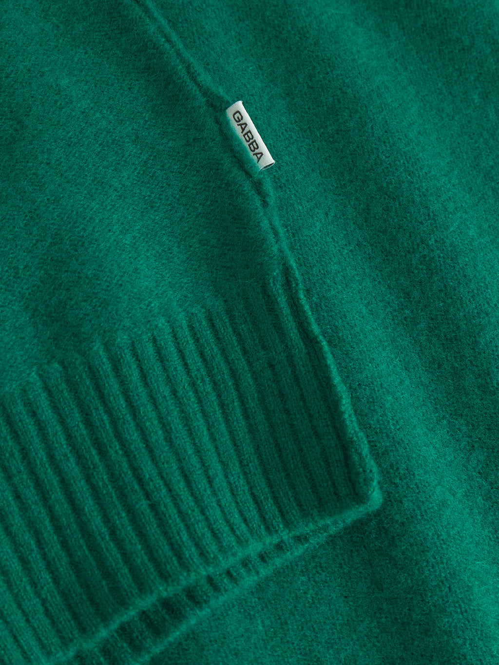 Close-up of the Tokota Aico LS - Green merino sweater by Gabba, showing a ribbed cuff and a small white fabric tag with the brand name. The detailed knitting texture is clearly visible.