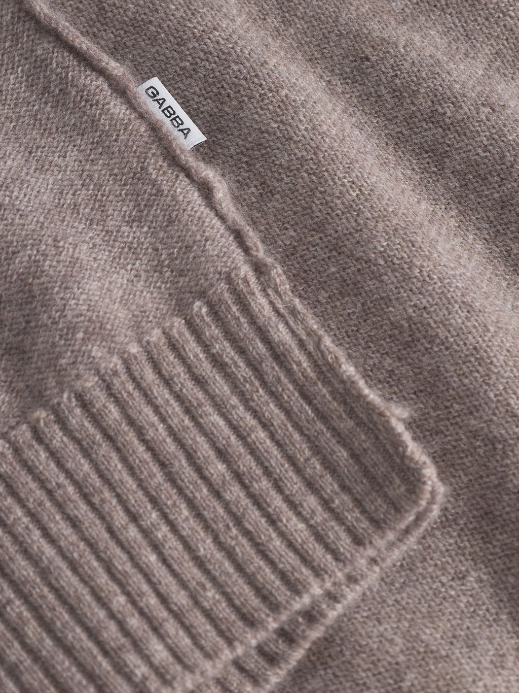 Close-up of the Tokota Aico LS sweater in light brown merino, featuring a ribbed cuff and a small white Gabba fabric label stitched near the seam.