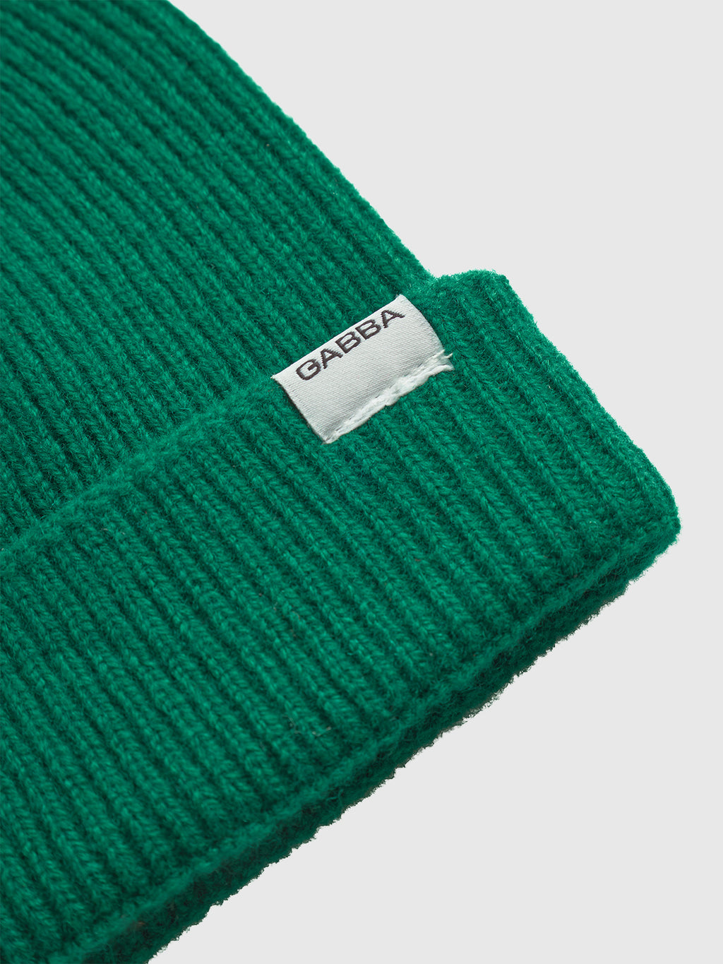 Close-up of the Biella Aico - Green ribbed knit beanie by Gabba, featuring a folded brim and a white tag with the brand name in black. Made from cozy merino wool, shown against a plain white background.