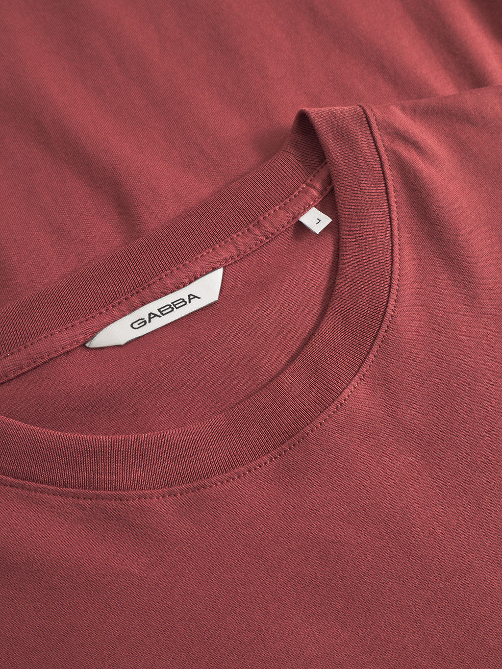 A close-up of the GABdune Logo GOTS SS T-Shirt in Red Russet, a classic fit by Gabba, shows a white collar label with the brand name and size A. The GOTS-certified fabric appears soft and features neat stitching.