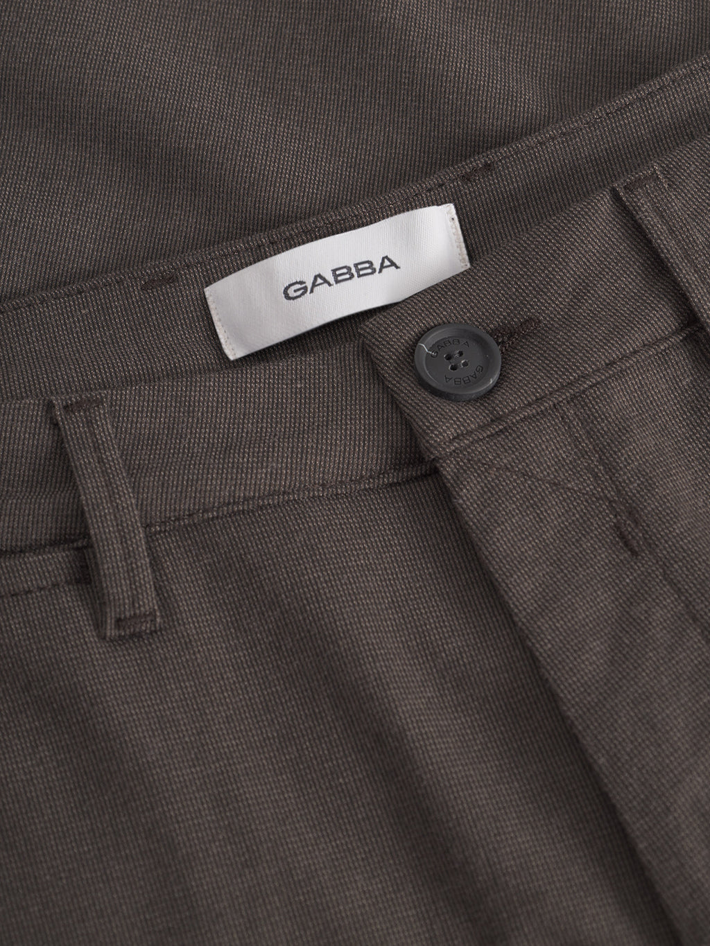 Close-up of the Paul Domo - Dk. Brown slim chino pant by Gabba, featuring textured brown fabric, branded buttons, belt loops, French pockets, and a white inner waistband label with the Gabba name.