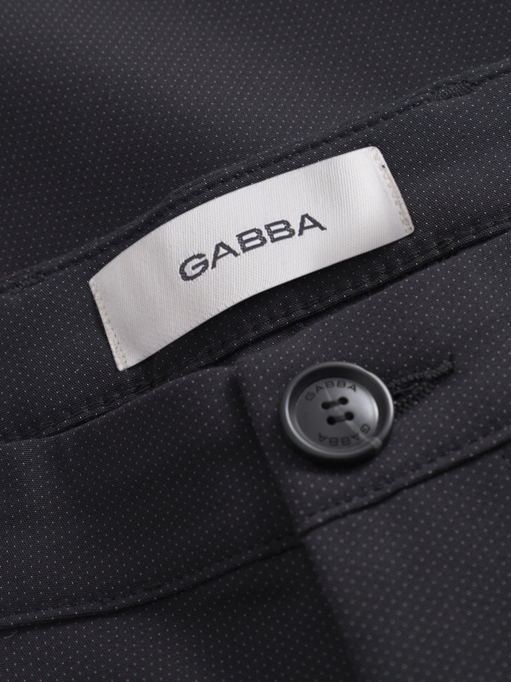 Close-up of Pisa Small Dot - Black pants by GABBA, showing black high-stretch cotton-blend fabric with tiny white dots, a white GABBA label, and a matching black button engraved with the brand on the pocket flap.