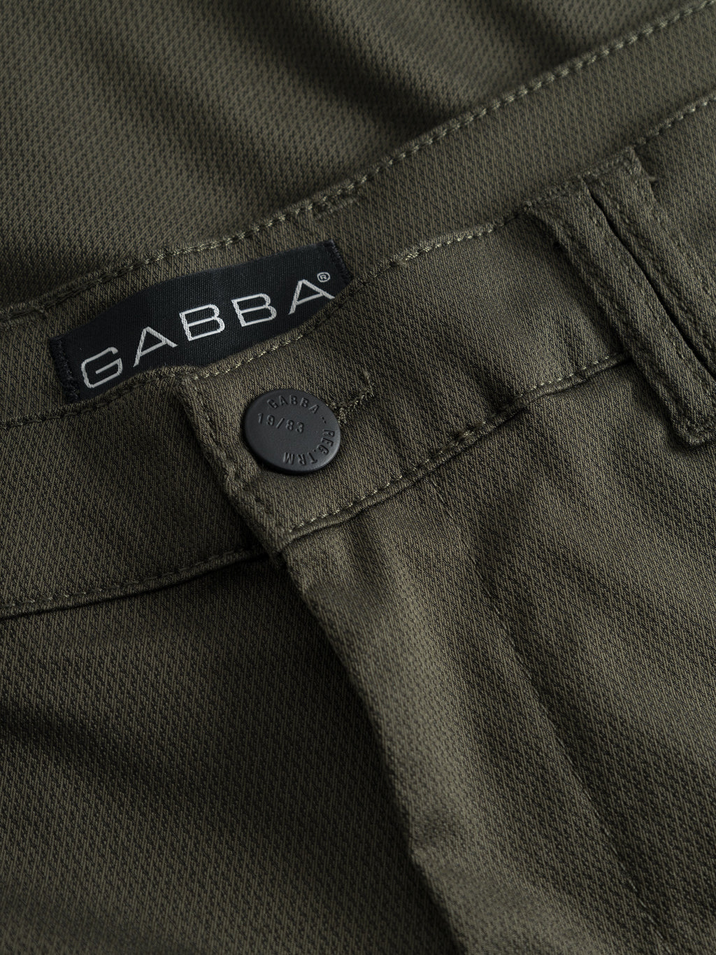 Close-up of GABBA Pisa Cargo K3280 Dale Pants - Army, featuring a black GABBA-stamped button, fabric tag by the waistband, visible stitching, super stretch fabric, and textured design for a comfortable slim tapered fit.