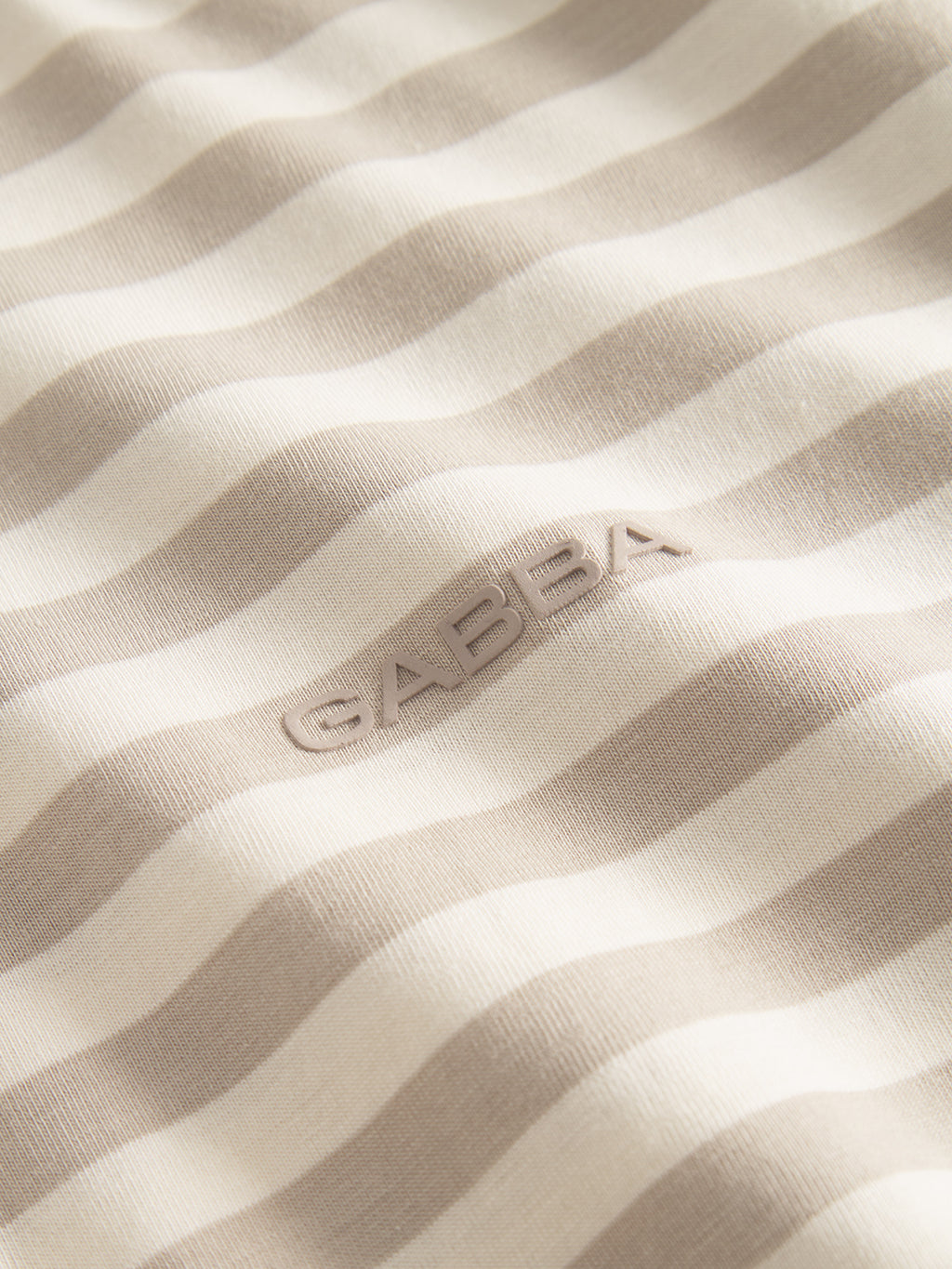 Close-up of the GABdune Logo Stripes SS GOTS t-shirt by Gabba, featuring beige and off-white horizontal stripes with “GABBA” embossed at the center. The texture and softness of this organic cotton tee are clearly visible.