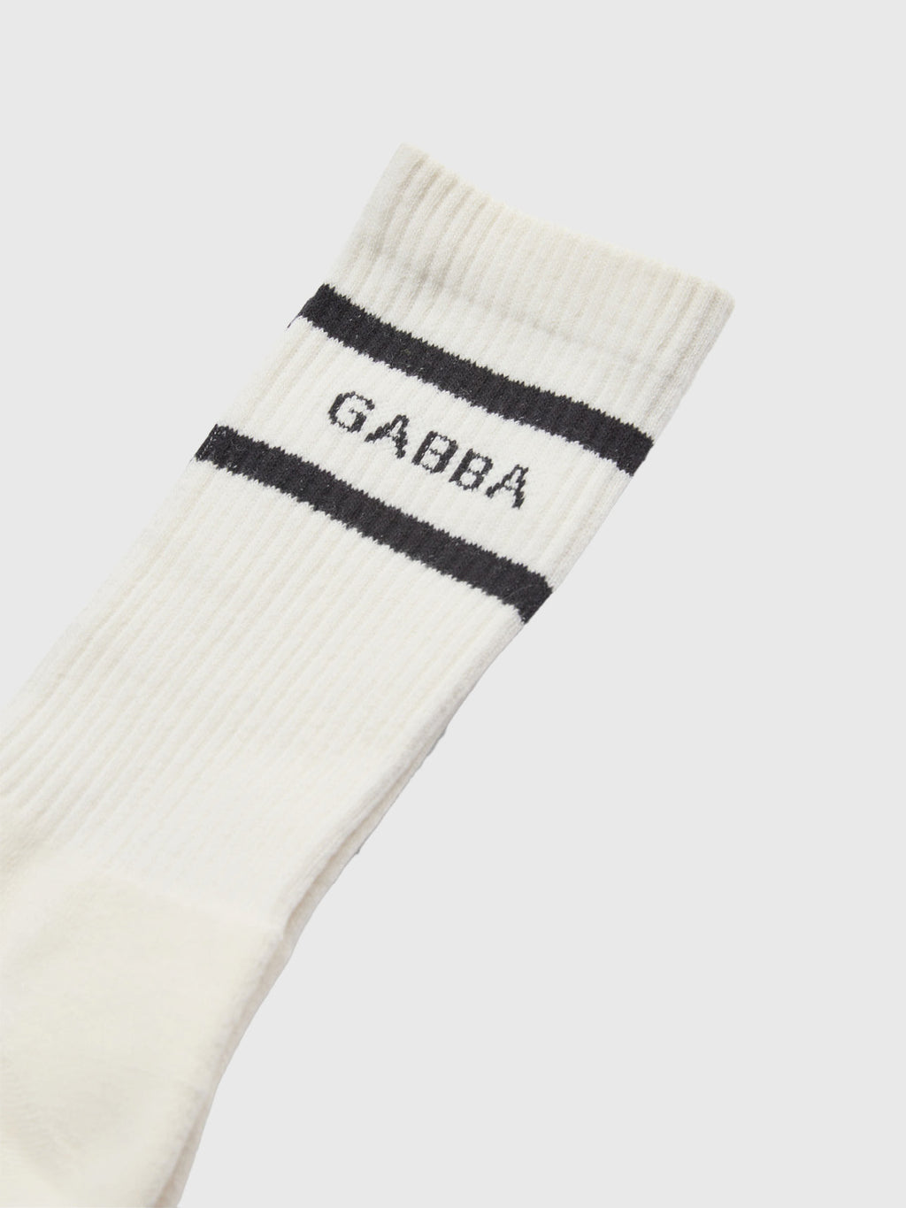 A close-up of GABBA's Loris Socks – White: a white ribbed sock with two black stripes near the top and GABBA between them, designed for daily wear and a comfortable fit.