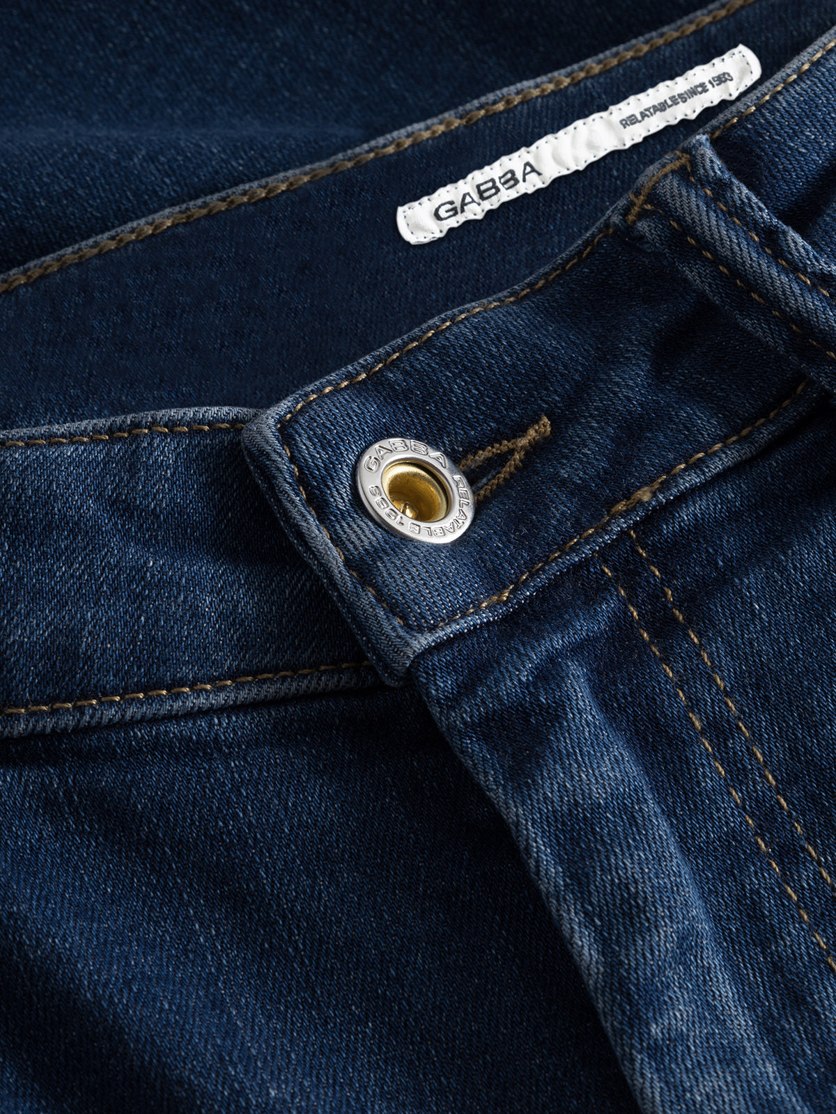 Close-up of GABjones K3870 slim jeans in mid blue denim by Gabba, highlighting the waistband, metal button with stitching, and a partial white Gabba label. Made from super-stretch fabric for comfort and style.