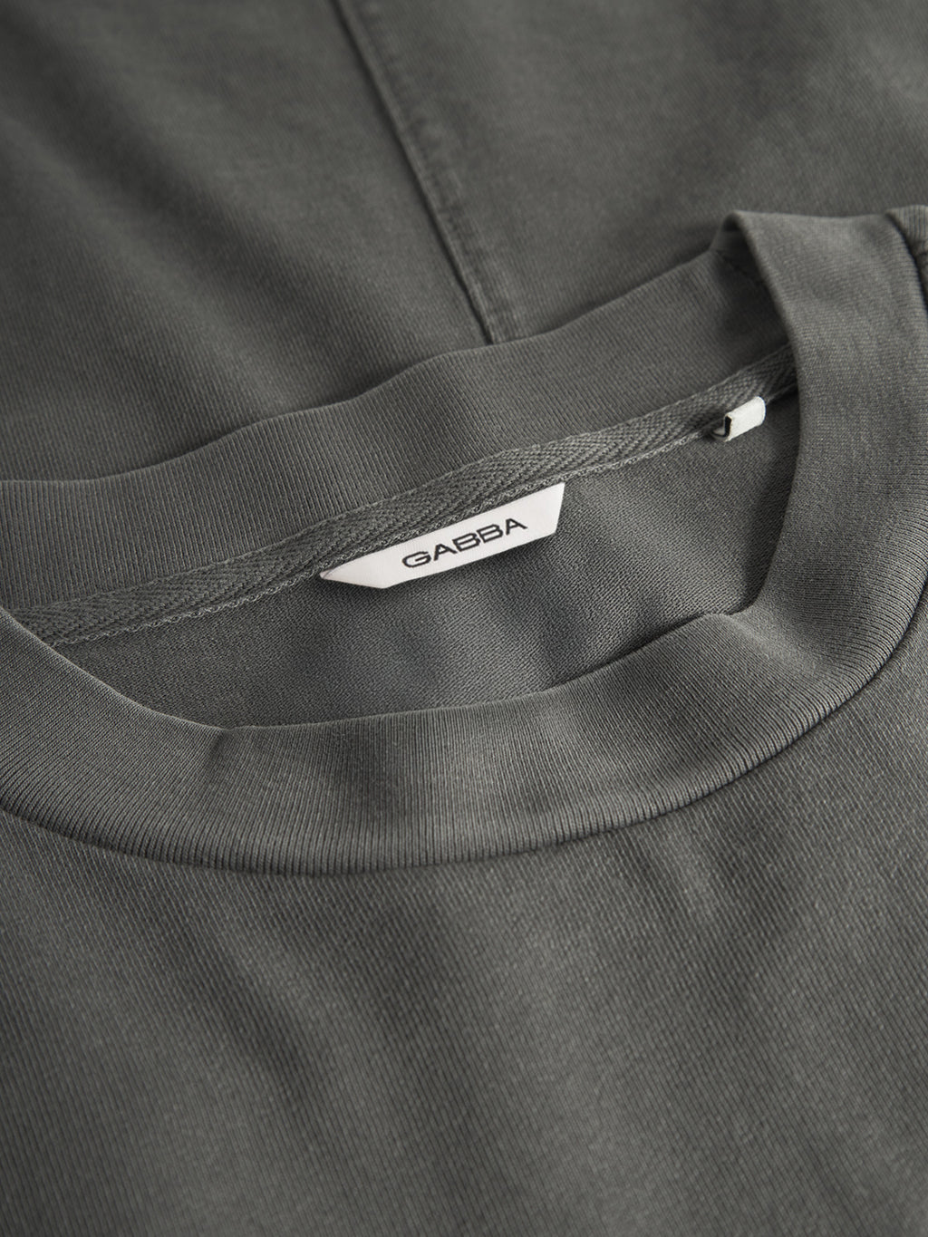 Close-up of the GABnigel Wix Boxy Hour SS t-shirt in dark grey by Gabba, heavyweight cotton with a crew neck. A white collar tag shows the Gabba brand name in black. The boxy tee is laid flat on a matching surface.