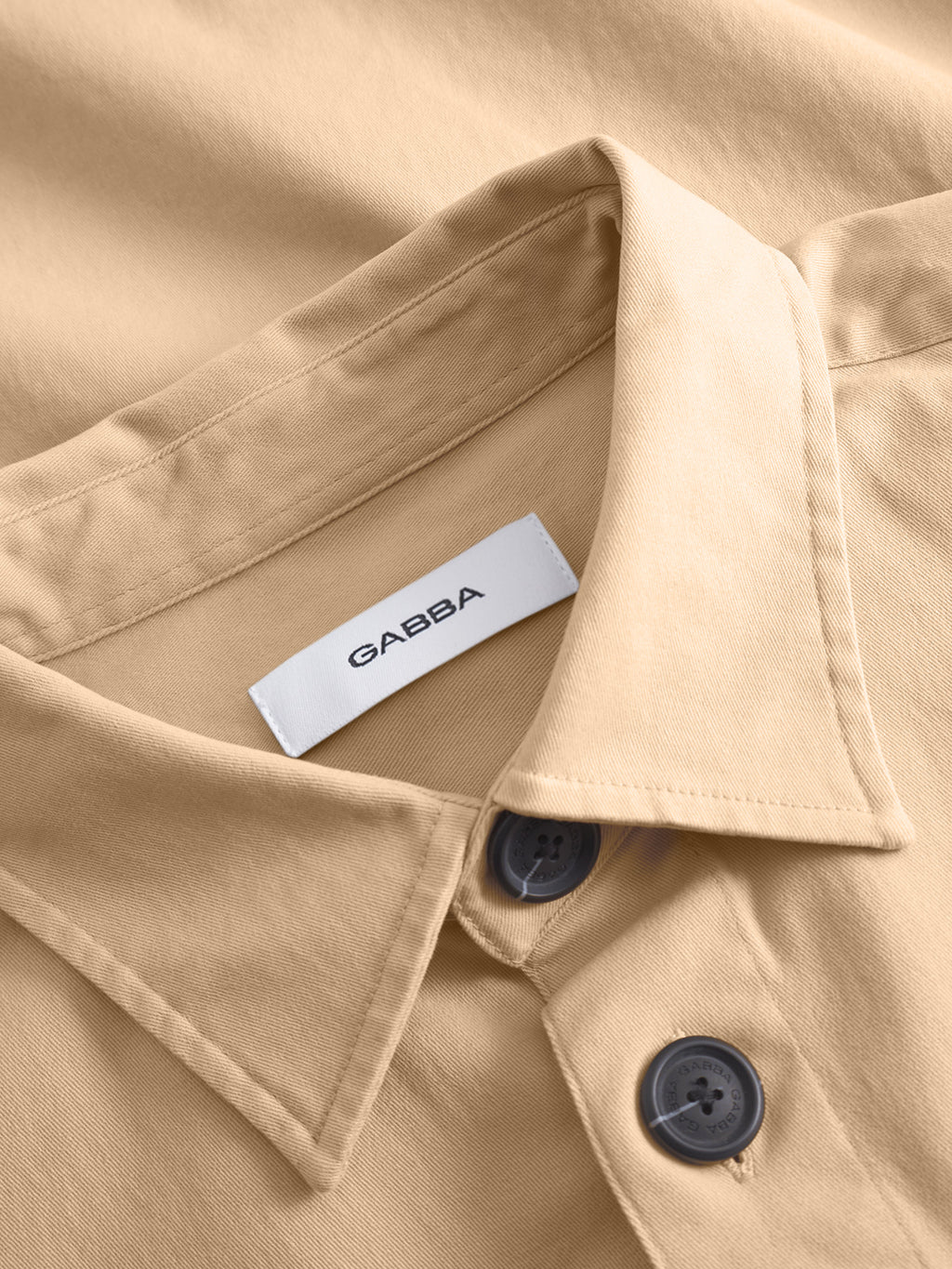 Close-up of the GABBA GABtopper LS Shirt in Humus, a beige collared overshirt with dark buttons, smooth fabric, neat stitching, chest pockets, and a white inner collar label showing 