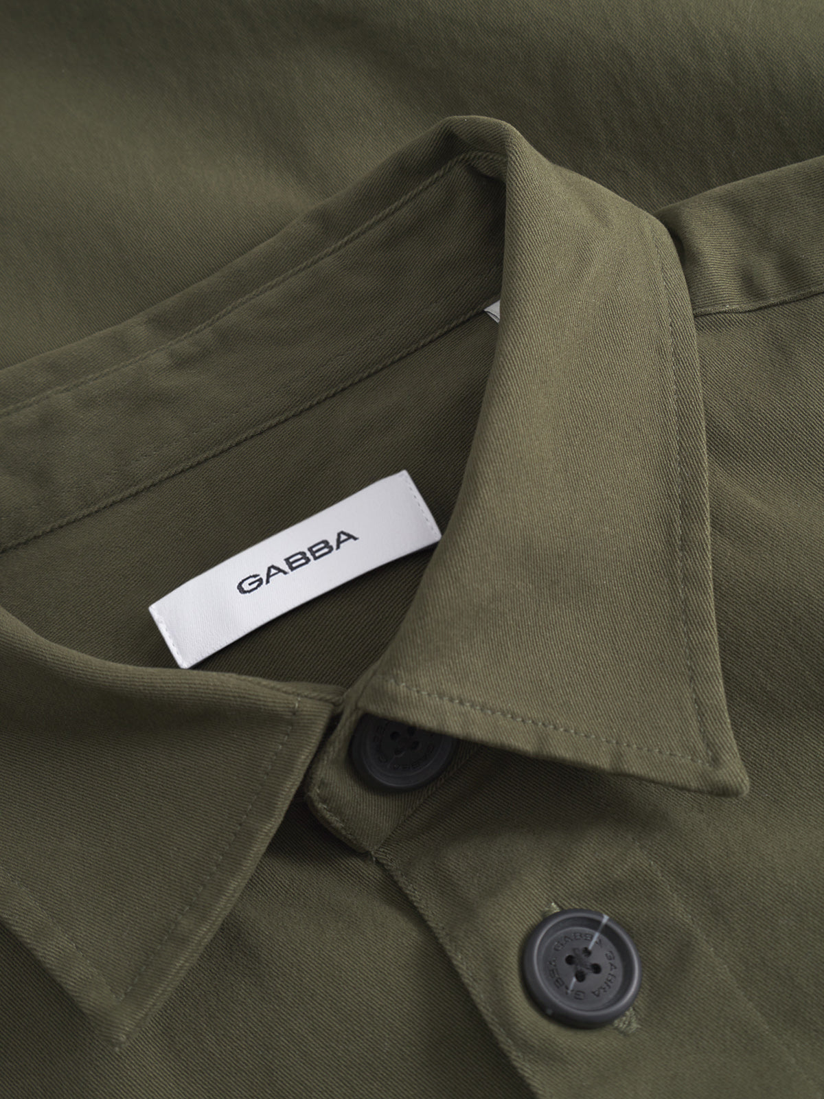 Close-up of the GABBA GABtopper LS Shirt in Army, featuring black buttons and a white tag with the GABBA brand name in black lettering inside the collar.