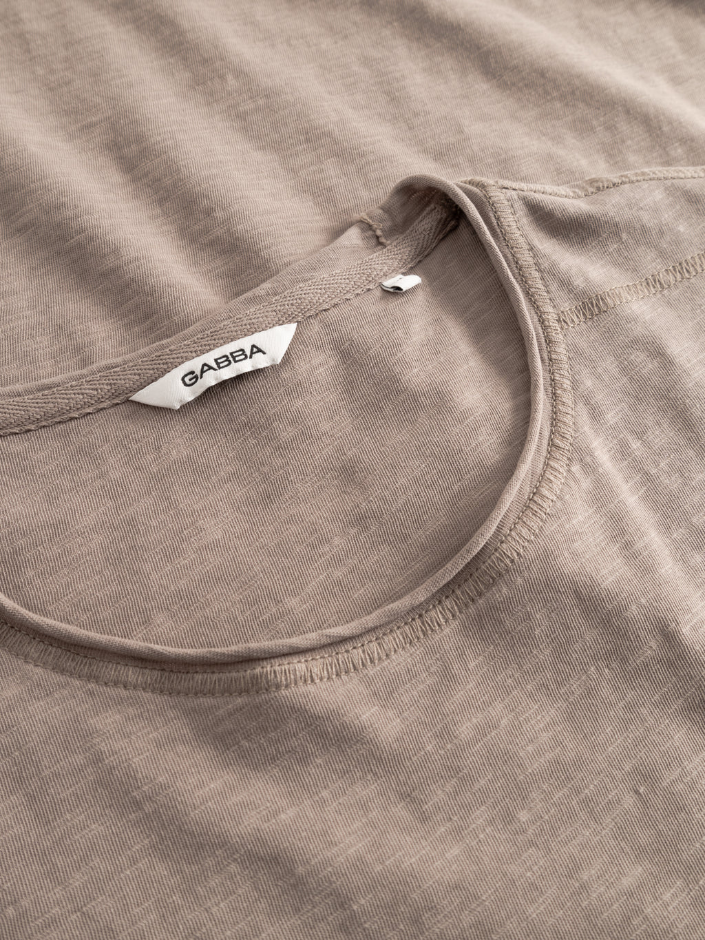 A close-up of the GABkostant New SS - Fungi T-shirt by Gabba in taupe-colored slub, semi-sheer 100% cotton, featuring neckline and stitching details, plus a white tag with 