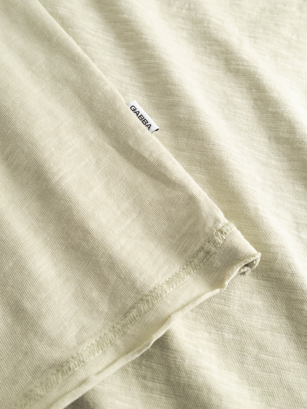 Close-up of the GABkostant New SS t-shirt by Gabba in Alfa Green, showcasing light beige slub fabric, a stitched hem, and a small white tag with Gabba branding in black.