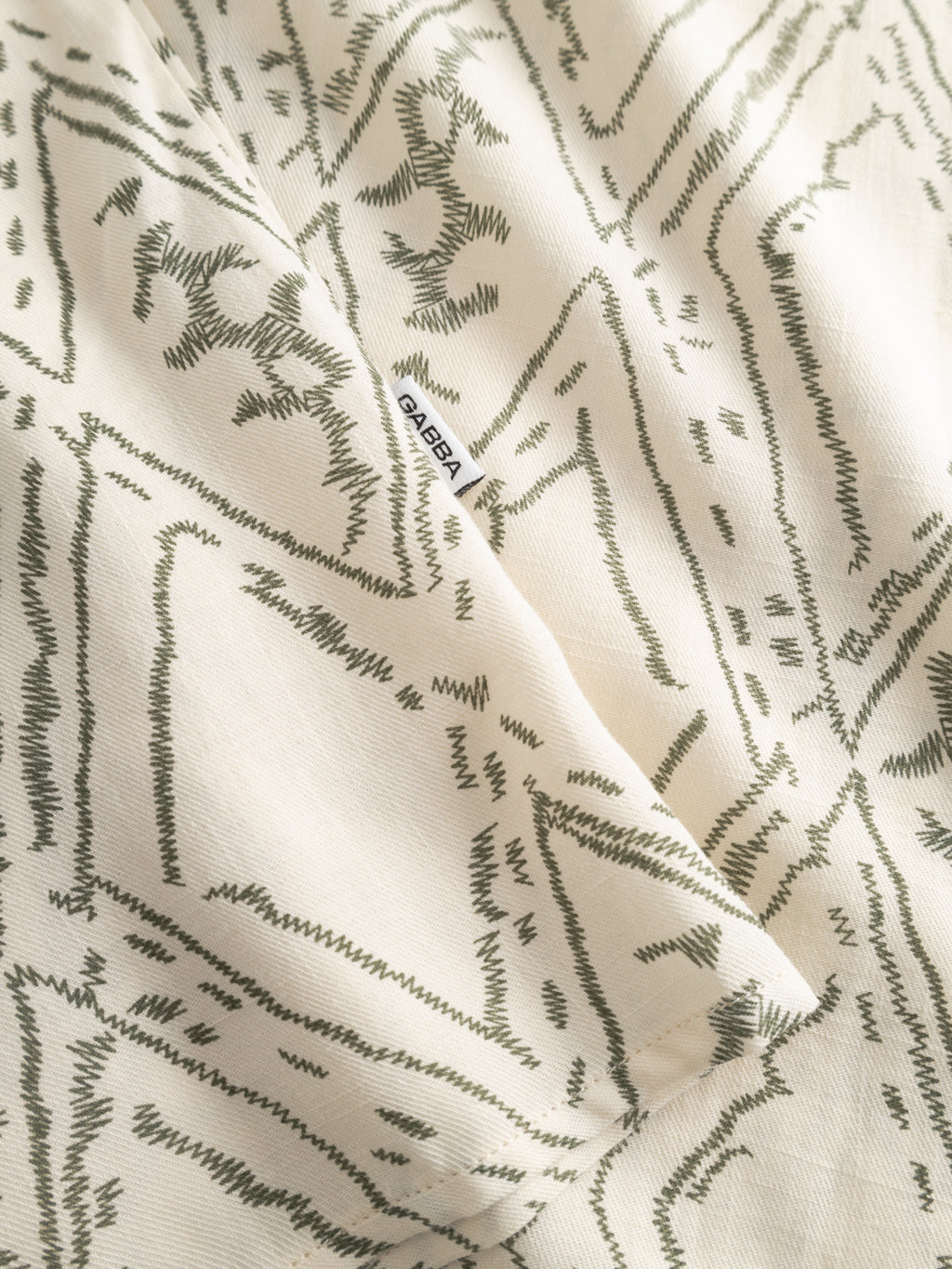 A close-up of cream-colored fabric from the Gabba GABsven Reg Zag SS Shirt - Multi Pattern shows abstract green zigzags and geometric designs, with a partially visible white OASIBA label along the seam.