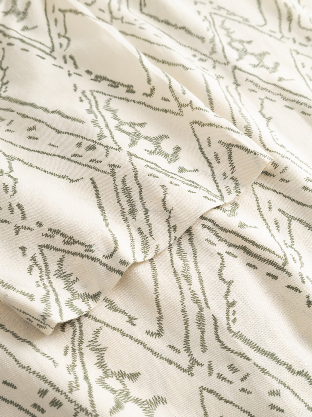 A close-up of cream-colored fabric from the Gabba GABsven Reg Zag SS Shirt - Multi Pattern, showcasing an abstract olive green angular geometric print. The soft material is gently folded, emphasizing its modern design and texture.