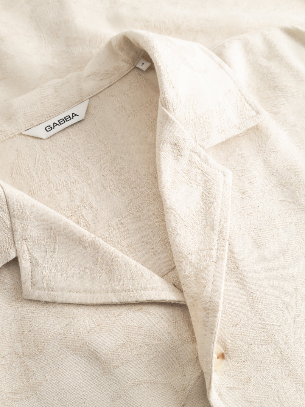 A close-up of the Gabba GABjens Over Resort Soft SS Shirt in ecru, an oversized linen blend with a notched collar, subtle all-over leaf print, and visible Gabba label inside the neckline.