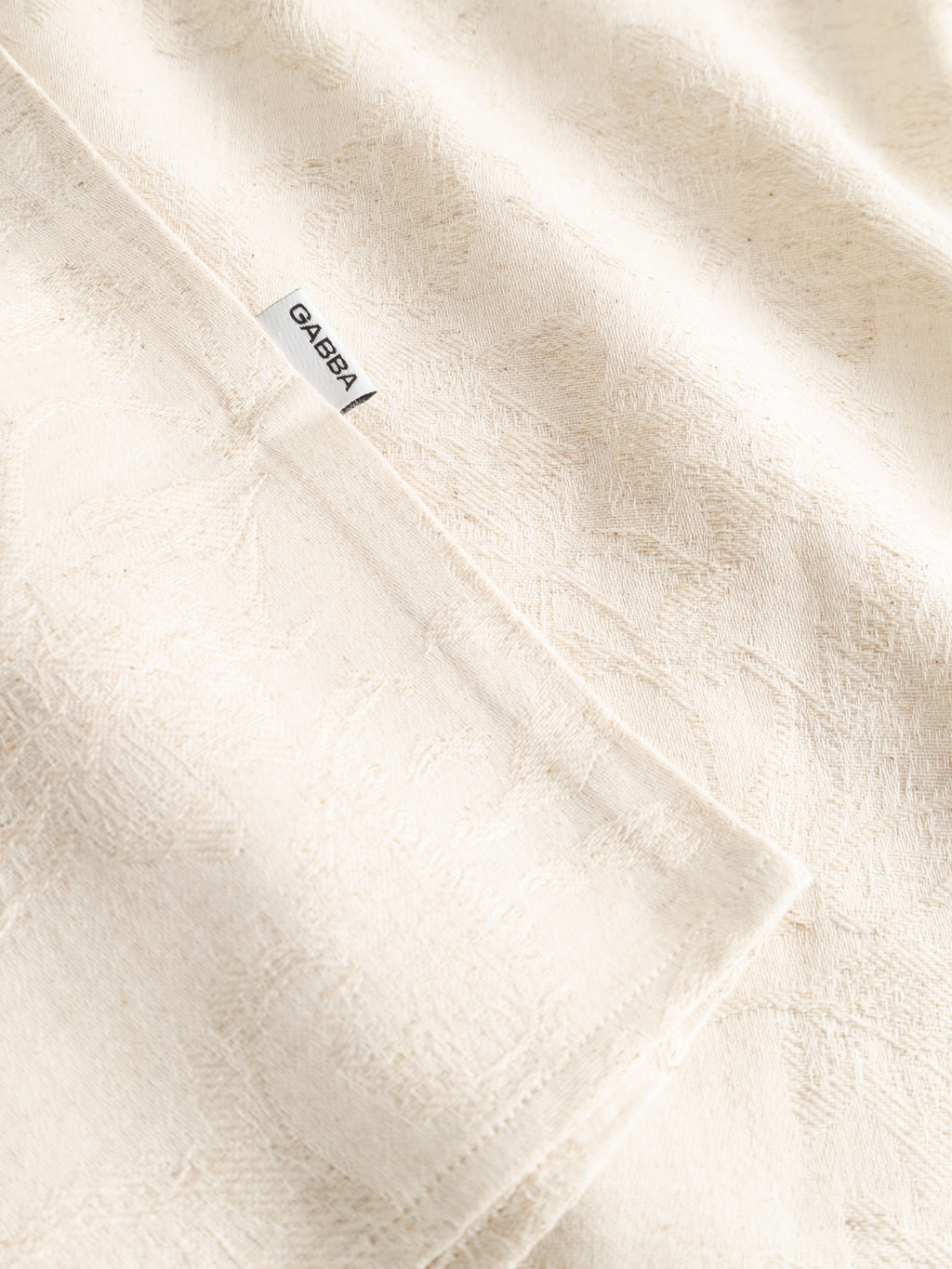 A close-up of textured ecru fabric from the Gabba GABjens Over Resort Soft SS Shirt, featuring a subtle pattern and a small white label with 
