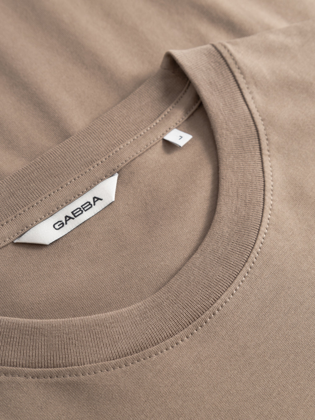 Close-up of the Gabba GABdune Logo GOTS SS T-Shirt in Fungi, an organic cotton tee with a visible white Gabba label and a small “Z” size tag near the collar. The taupe fabric looks soft and smooth.