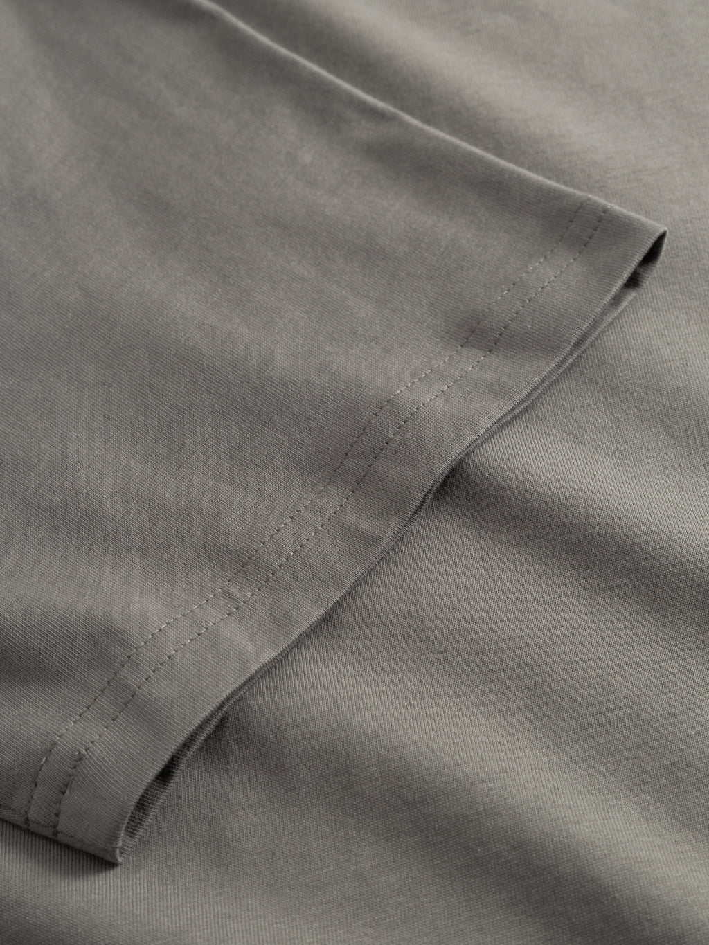 A close-up of the GABdune Logo GOTS SS T-Shirt in Castor Gray by Gabba, highlighting the organic cotton hem and stitching on a folded sleeve edge, with a smooth, soft texture.