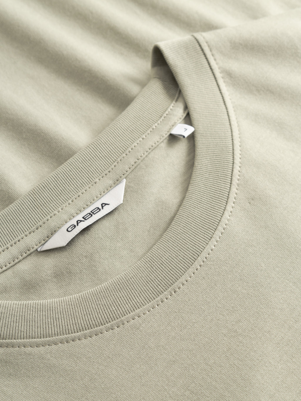 A close-up of a light green GABdune Logo GOTS SS T-Shirt by Gabba features the collar, detailed stitching, and tags, including a visible Gabba brand label and a small size tag marked 7.