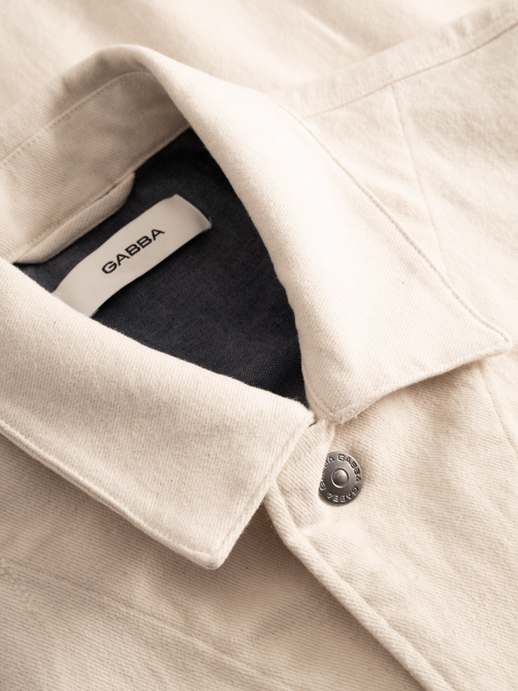 Close-up of the Gabba GABbeta Twill - Ecru jacket, featuring a light beige hue, pointed collar, metal snap button, and a visible black GABBA label inside the collar.