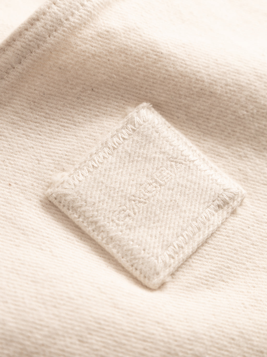 Close-up of Gabba's GABbeta Twill - Ecru fabric, showing its textured, off-white finish and a square patch with visible stitching and subtle central text, evoking classic workwear overshirts.