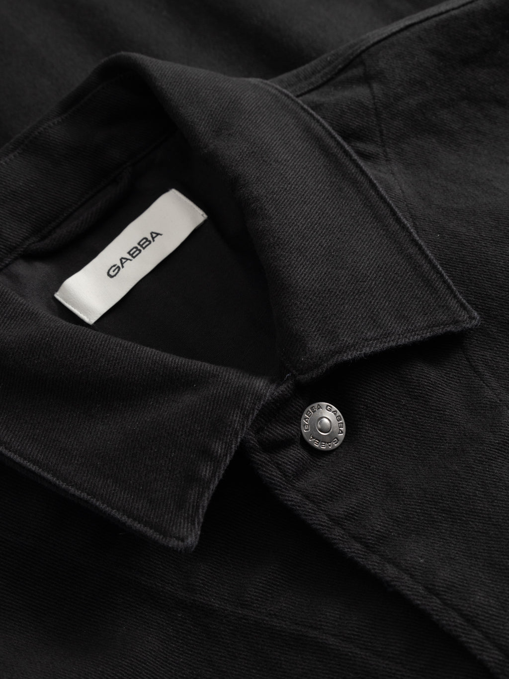 Close-up of the Gabba GABbeta Twill - Black shirt featuring garment-dyed black twill, a metal button, and a white Gabba label inside the collar. The textured fabric highlights its quality and comfort.