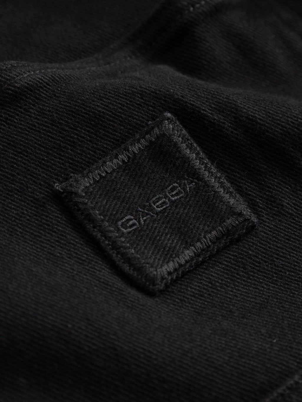 Close-up of GABbeta Twill - Black by Gabba, showing black garment-dyed twill fabric with a square stitched patch and GABBA embroidered in black—ideal for a comfortable shirt or workwear overshirt.
