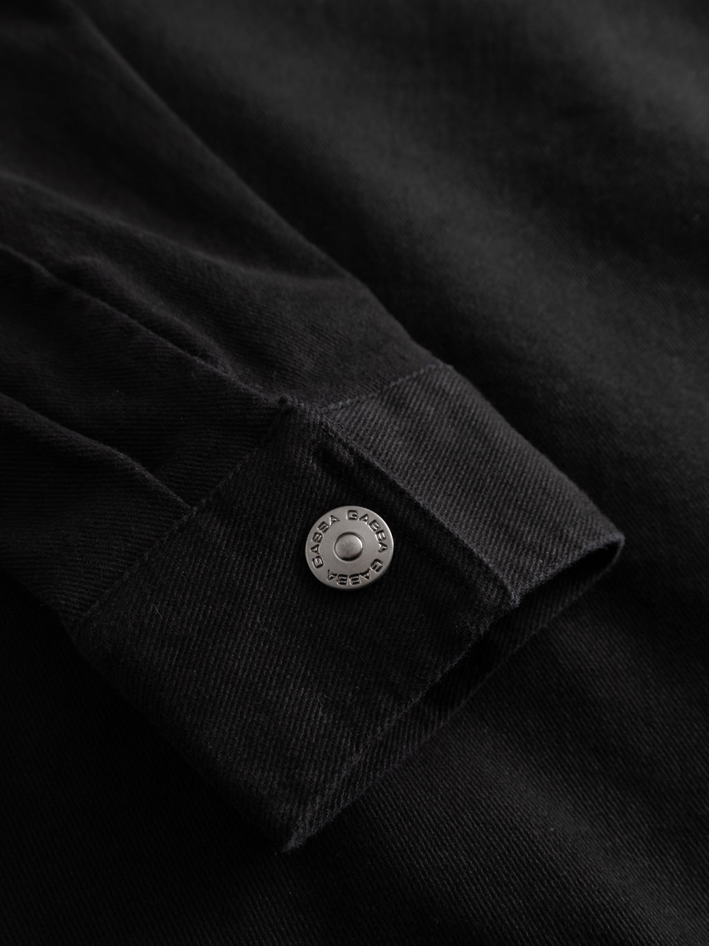 Close-up of the GABbeta Twill - Black workwear overshirt by Gabba, showing its textured black fabric cuff with a metallic button engraved 