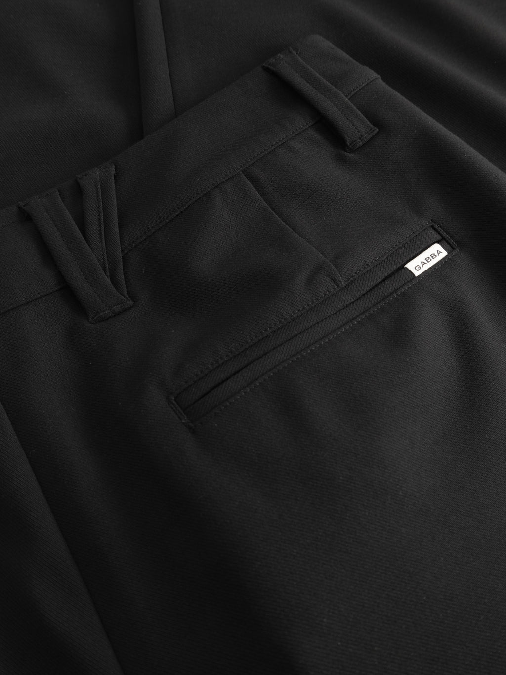 Close-up of the back pocket and waistband of Gabba GABfirenze Phin Cropped GRS - Black trousers, featuring a black zipper, a small white tag with text, and smooth, neatly stitched fabric.