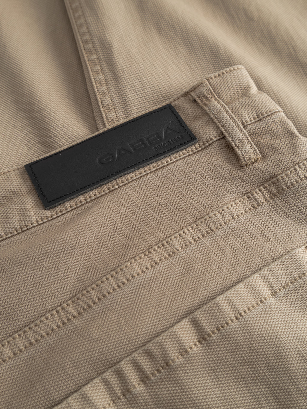 Close-up of beige textured GABmarc 5poc Marlo - Coriander regular-fit jeans by Gabba, showing a black leather patch label with the brand name embossed next to a belt loop and seam details.