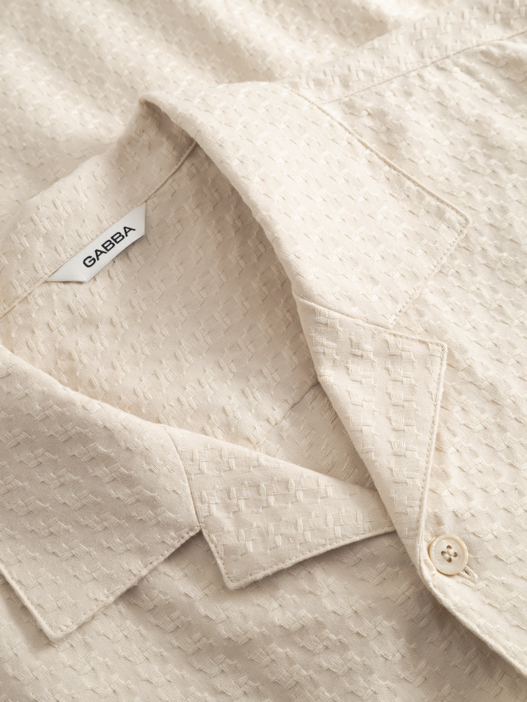 Close-up of the GABtimo Resort Neal SS Shirt in Birch by Gabba—a beige shirt with a pointed collar, buttoned front, visible Gabba label, and textured jacquard 3D motif.