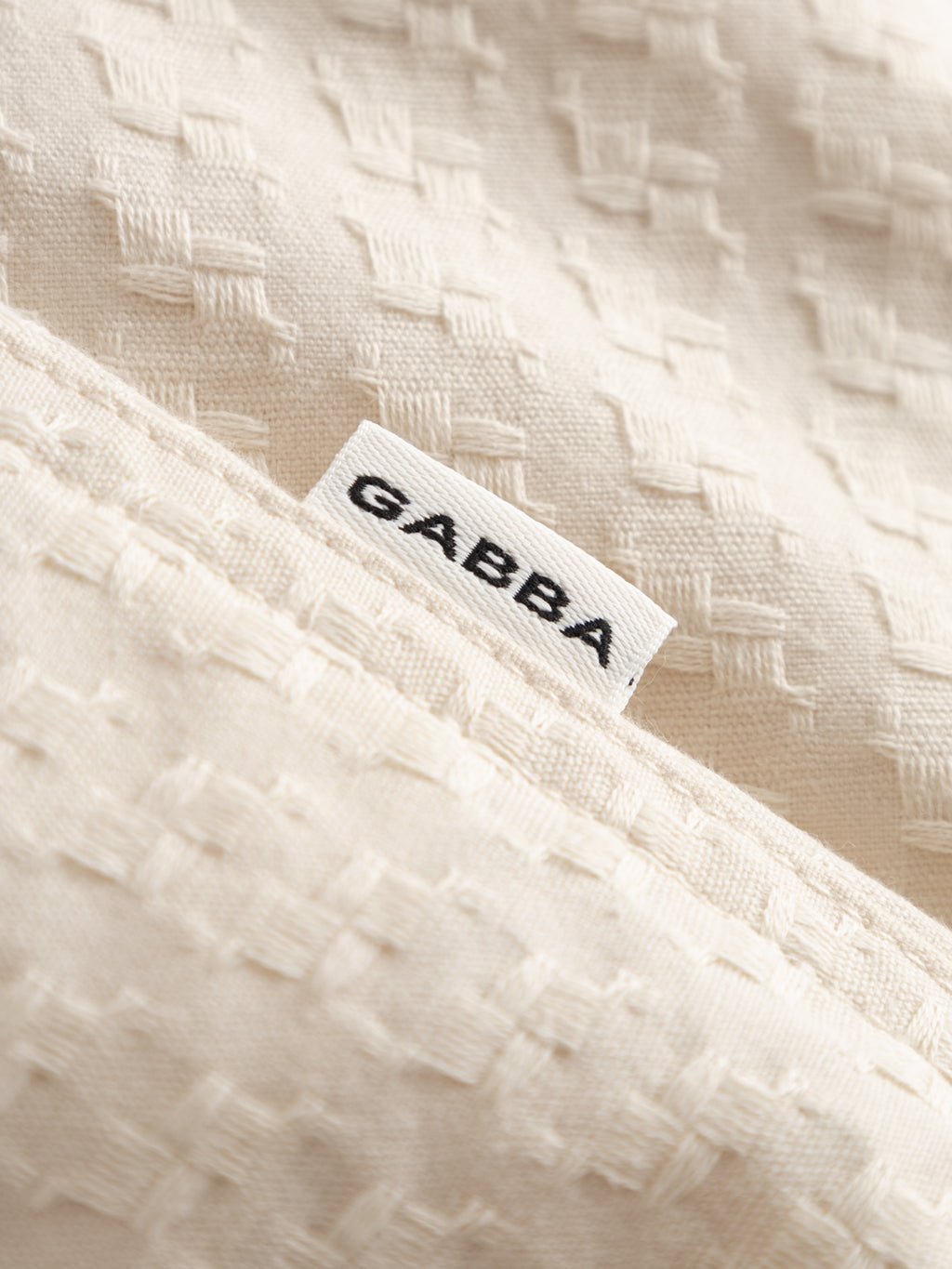 A close-up of the cream-colored GABtimo Resort Neal SS Shirt - Birch by Gabba, featuring textured jacquard fabric and a woven 3D motif, displays a white label with 