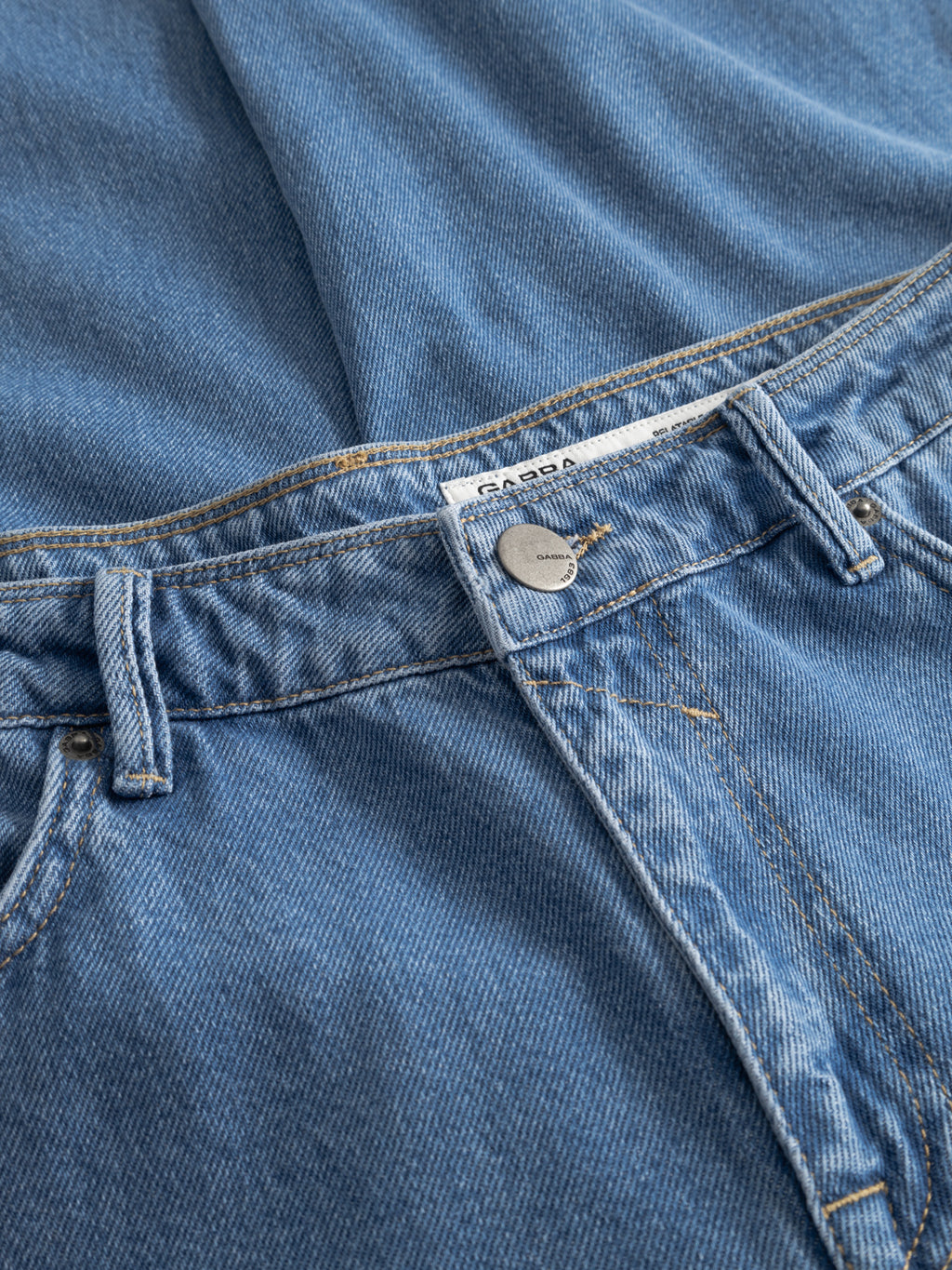 A close-up of Gabba’s GABmax 5poc Mid Jeans in mid blue denim shows the waistband, classic 5-pocket styling, metal button, belt loops, visible stitching, and part of a matching denim top.