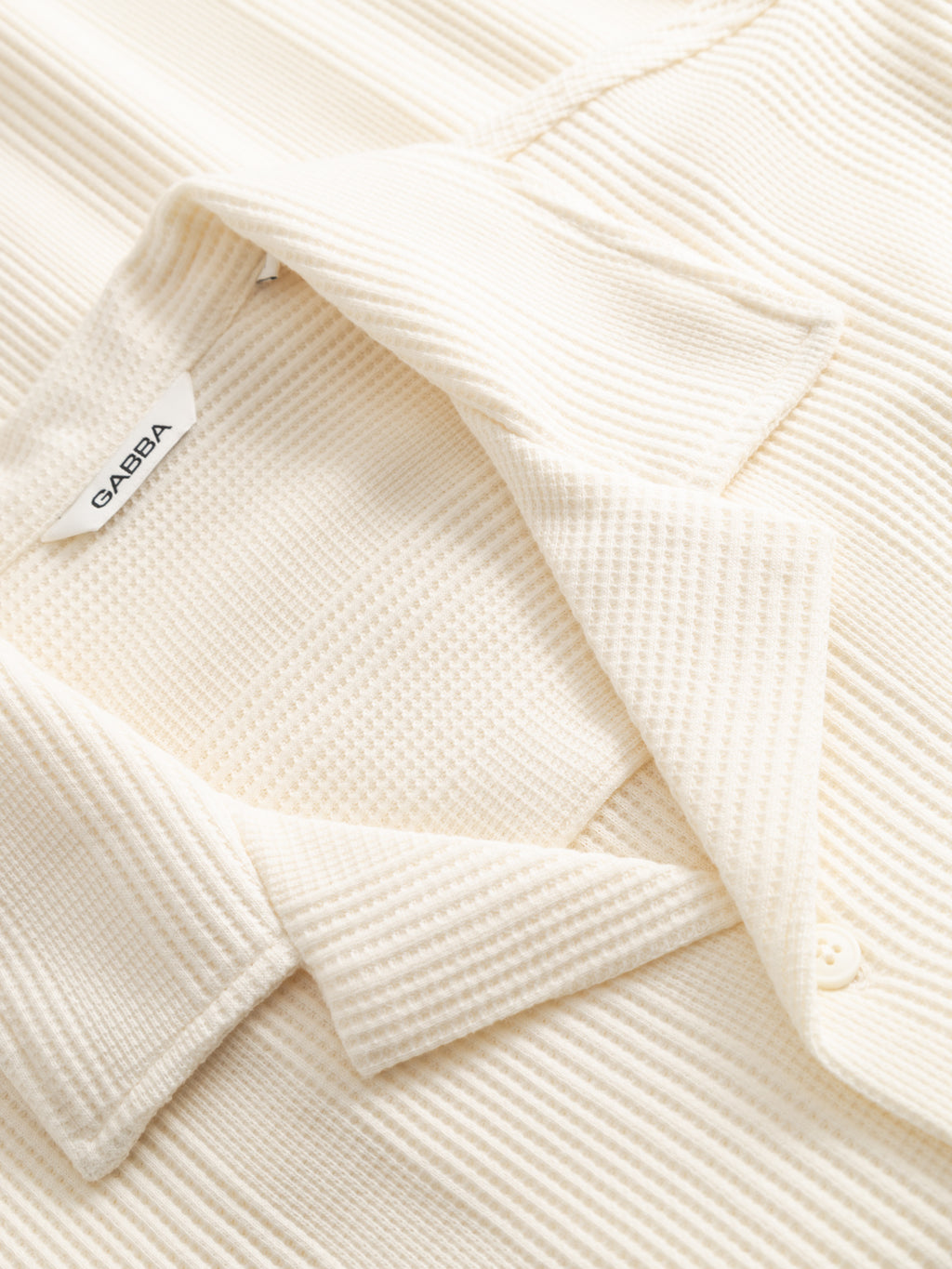 Close-up of a bone white GABvalente Wup SS T-Shirt by Gabba, featuring ribbed knit fabric, relaxed fit, open resort collar, and a white GABIA tag. A button is partially visible at the bottom right.