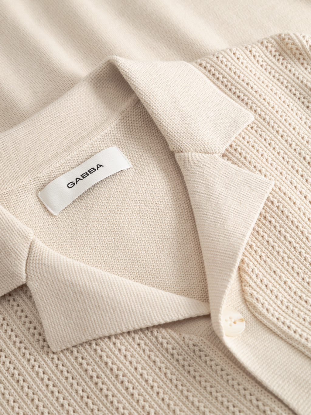 A close-up of the GABgeno Resort Bride SS Knit - Birch by Gabba, a beige cable knit shirt with a folded collar, button, and textured breathable cotton, featuring a white label that reads Gabba.