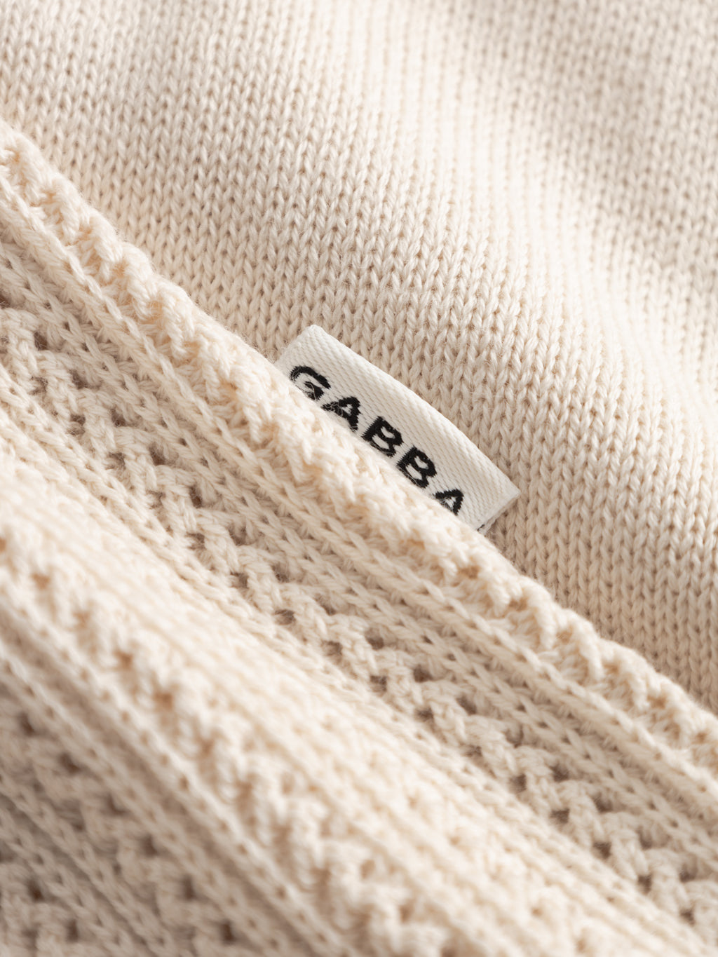 A close-up of the GABgeno Resort Bride SS Knit - Birch by Gabba in breathable cream cotton, showing a small white label with Gabba’s logo and highlighting the garment's smooth and textured knit patterns.