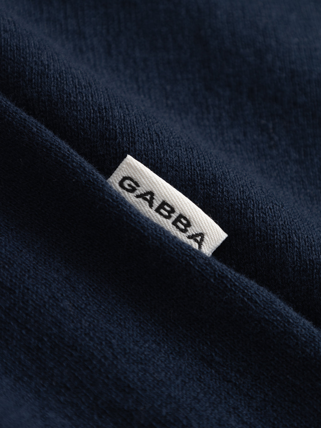 A close-up of navy GABlima Cot LS Knit by Gabba, made from 100% cotton, shows a small white Gabba label partially visible between two fabric folds.