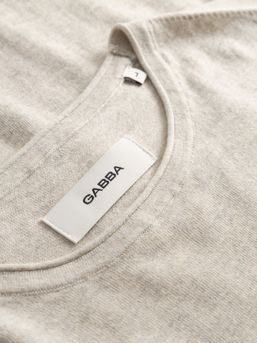 Close-up of the neckline of a sand melange GABlima Cot LS Knit by Gabba, showing seamless detailing, a white fabric tag with the brand name Gabba in black, and an additional size 1 label.