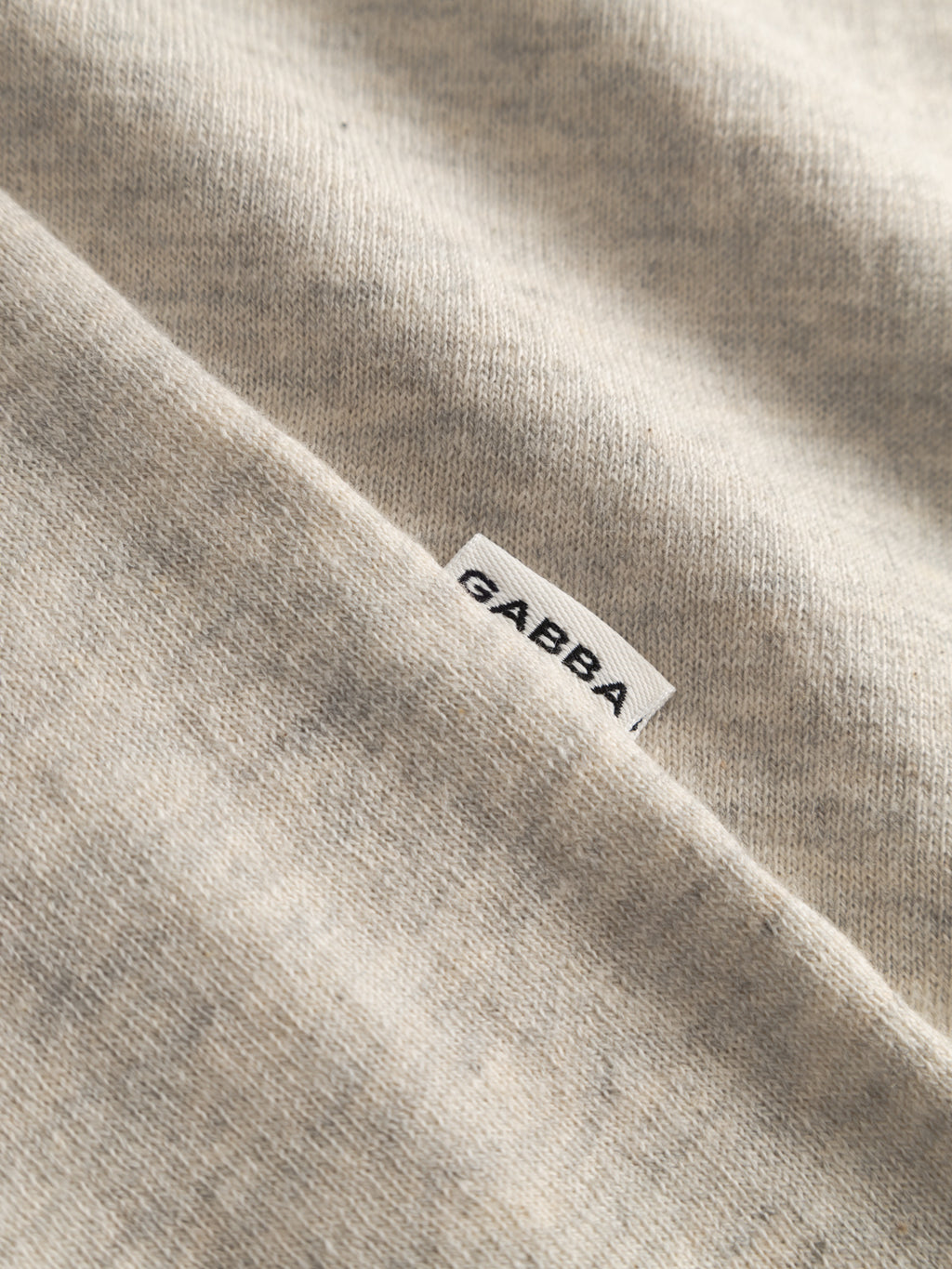 A close-up of the GABlima Cot LS Knit - Sand Mel. by Gabba shows light beige, 100% cotton fabric with a soft, textured finish and seamless detailing, plus a partially visible white tag displaying the Gabba brand.