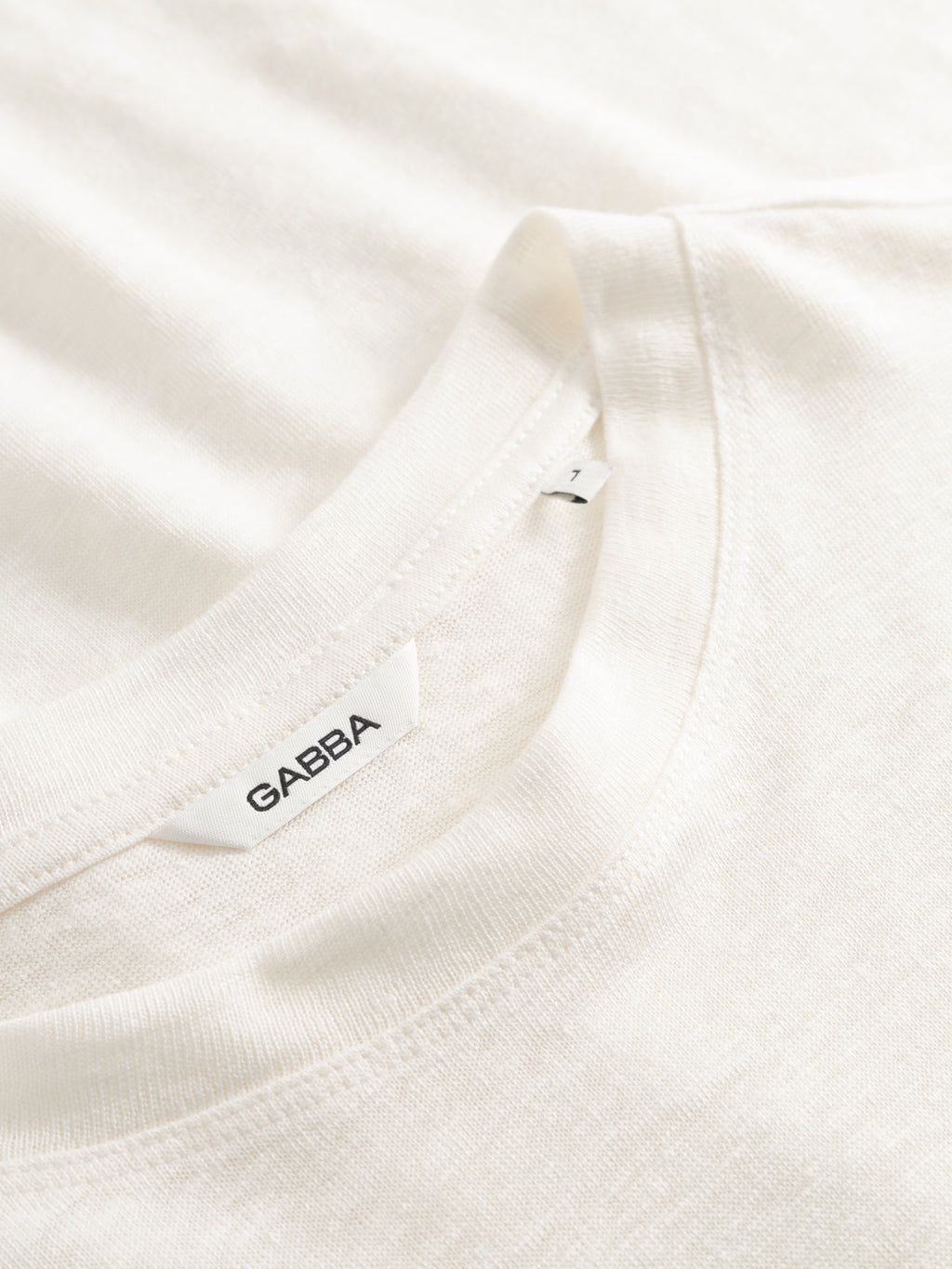 Close-up of the GABdune Linen SS T-Shirt - Birch by Gabba, showing its white linen texture and neckline tag with the Gabba logo. The regular fit shirt is laid flat on a matching white surface.