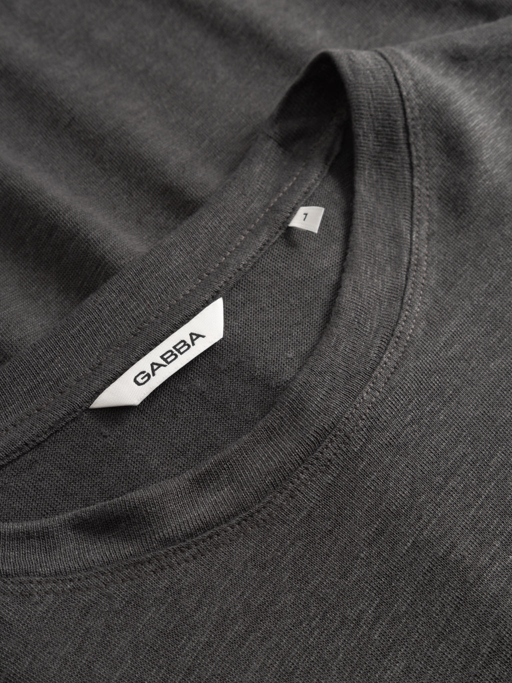 Close-up of the GABdune Linen SS T-Shirt in Pirate Black, a dark gray regular fit tee from Gabba. The image highlights the collar, a white label featuring the Gabba logo in black text, and a small size tag marked with the number 1. The linen-blend fabric appears soft and subtly textured.