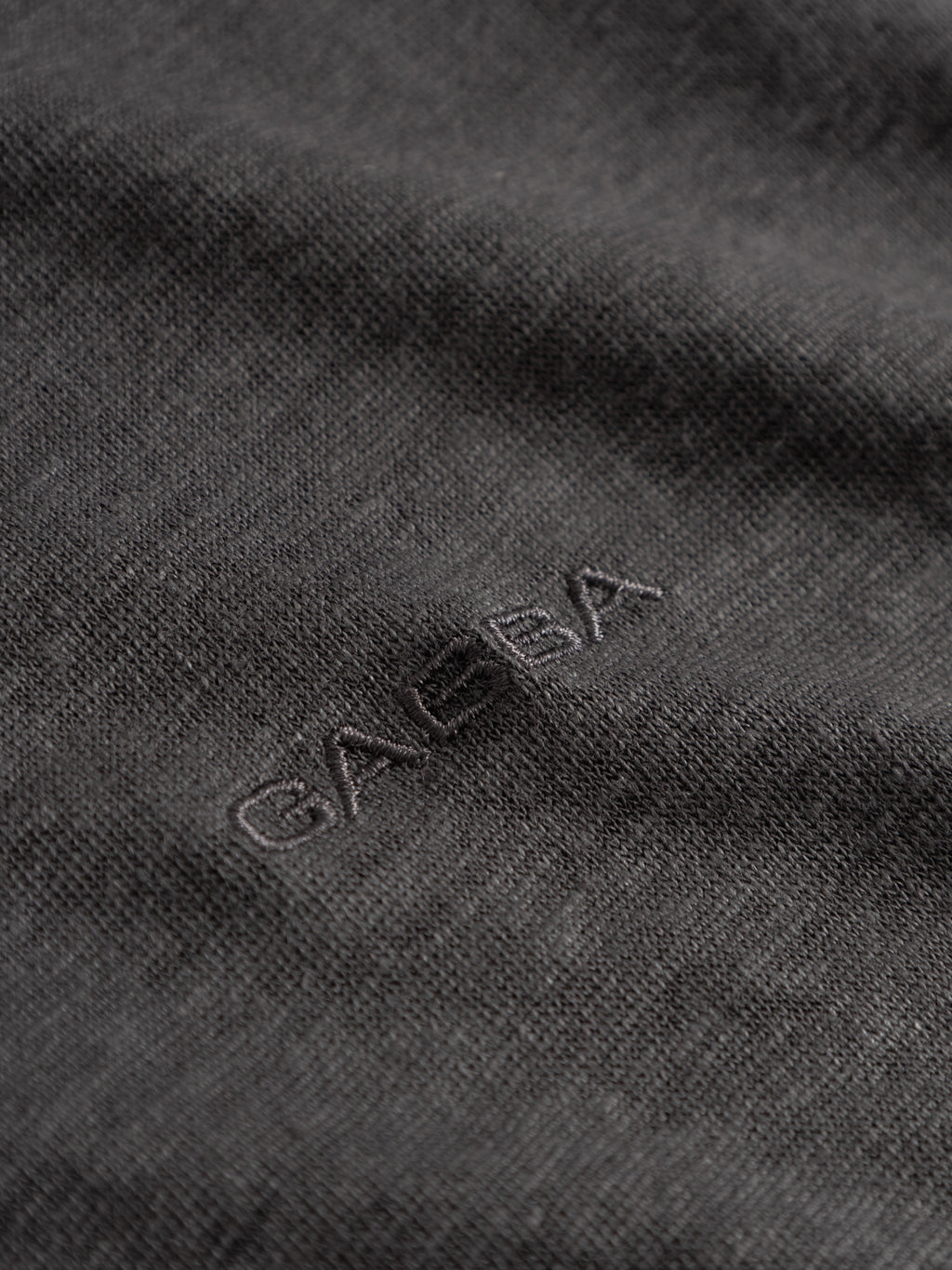 Close-up of the dark, textured fabric from the GABdune Linen SS T-Shirt in Pirate Black by Gabba, featuring “GAEBA” subtly embroidered in matching thread near the center— a refined nod to the iconic Gabba logo.