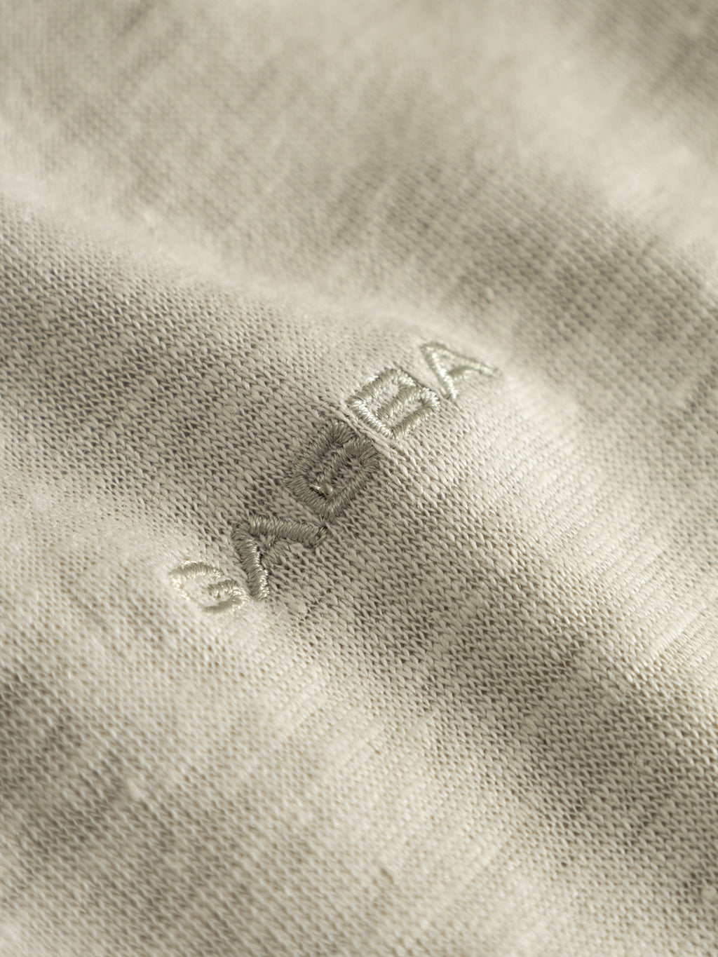 Close-up of the GABdune Polo Linen SS T-Shirt in Mineral Gray by Gabba, featuring textured light beige fabric with a subtle embroidered logo in matching thread and stitching reminiscent of a refined linen polo.