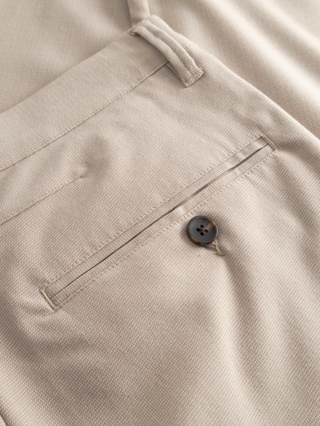 Close-up of the back pocket on Gabba’s GABpaul Deme Pant in Birch: beige slim-fit trousers with a dark button closure, visible belt loop, and subtly textured micro-patterned fabric.