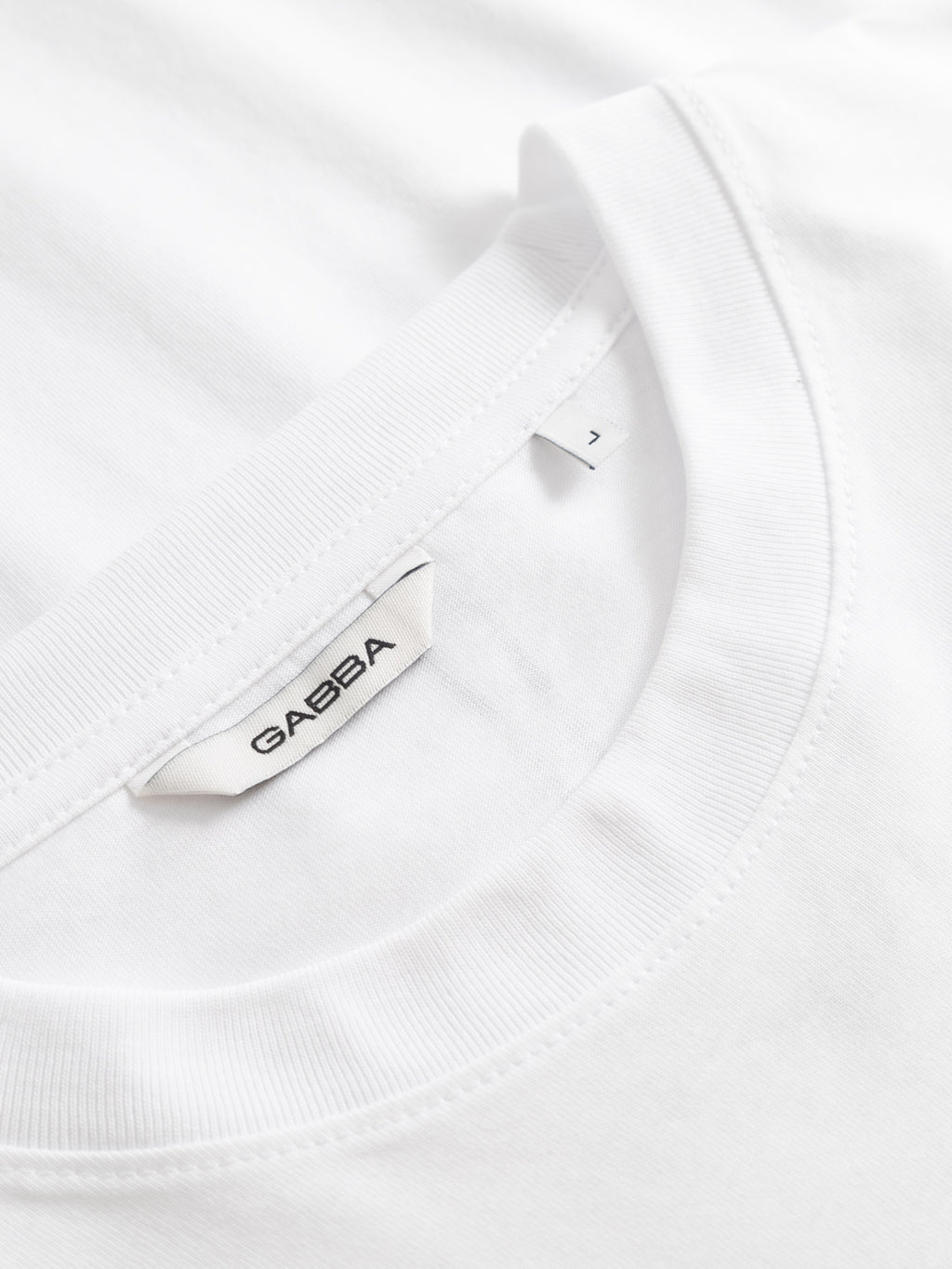 Close-up of a white GABnigel NBH SS T-Shirt GOTS showing the collar and a white tag featuring the Gabba brand name in black letters.