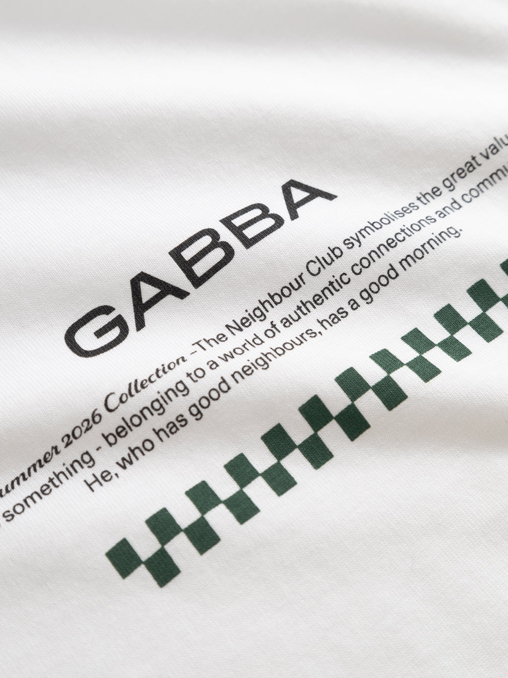 Close-up of the Gabba GABnigel NBH SS T-Shirt GOTS in white, featuring bold black “GABBA” lettering, neighbor and community-themed text, and a green checkered pattern at the bottom.