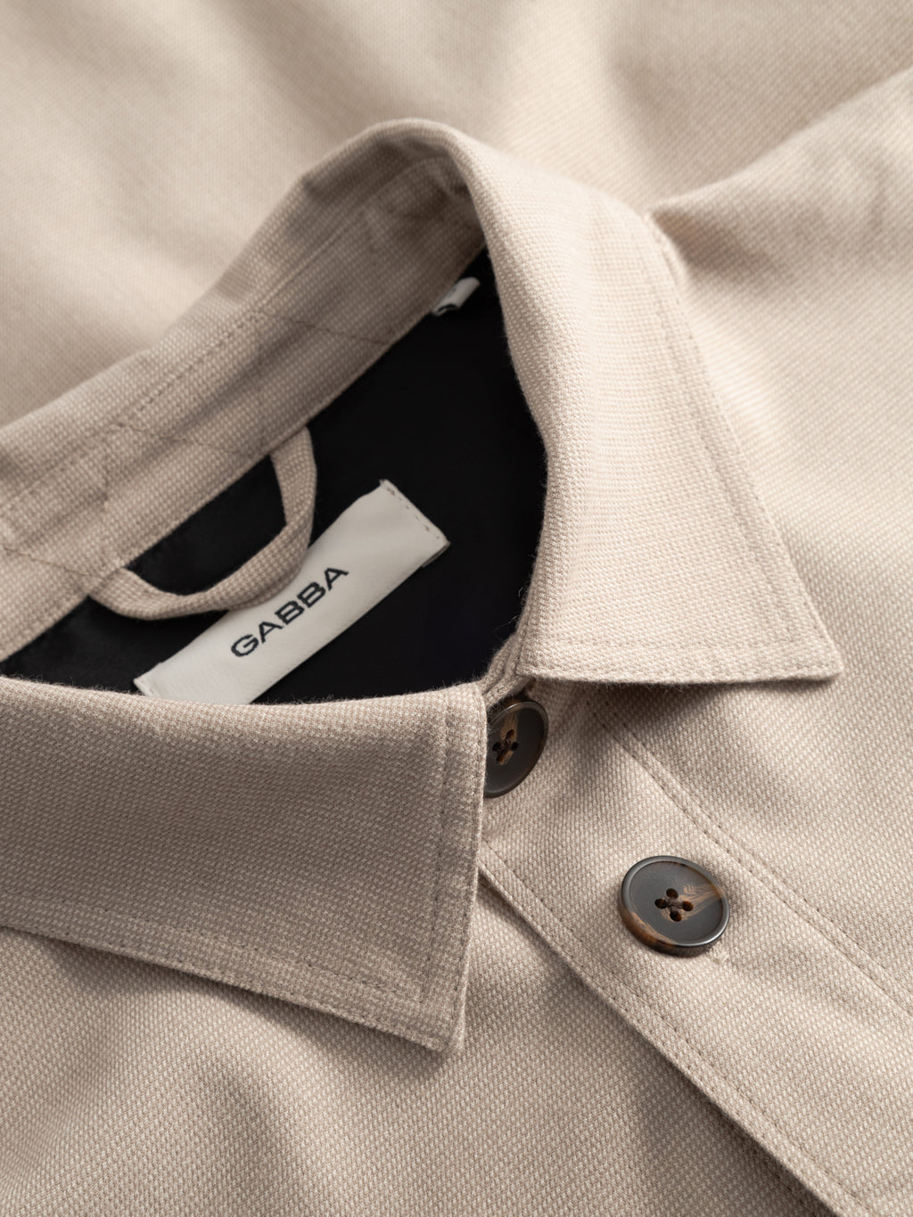 Close-up of the GABtogger Base Deme LS Overshirt in Birch by Gabba—beige, textured, with dark buttons, a collar, and a white neck label reading Gabba—a stylish classic for any occasion.